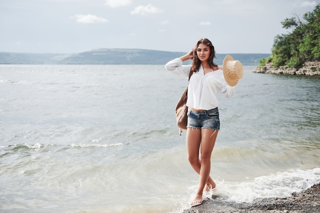 모자와 배낭에 아름 다운 젊은 여자는 장난스럽게 물에 의해 걷는다. 따뜻한 여름날은 자연의 모험과 모험을위한 좋은 시간입니다.