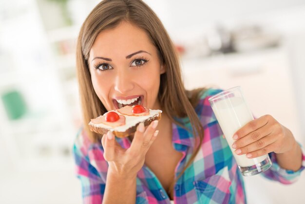 Beautiful young woman having breakfast in the domestic kitchen. She is eating fresh sandwich and drinking milk. Looking at camera.