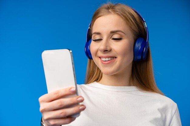 青い背景でスマートフォンで音楽を聴いているヘッドフォンをかぶった美しい若い女性