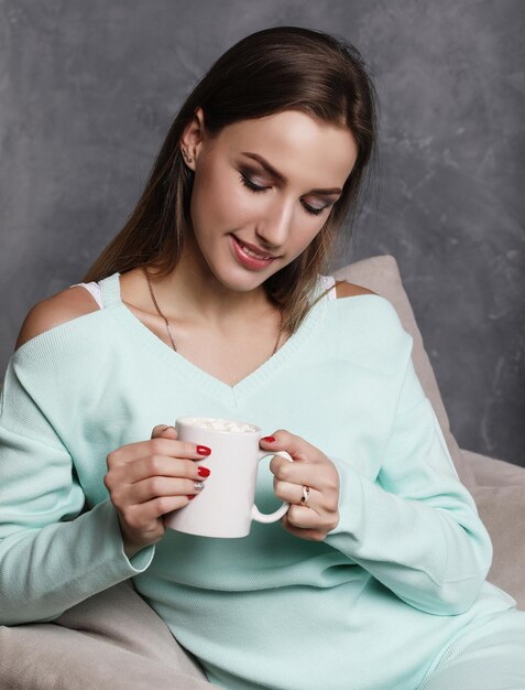 Beautiful young woman holding coffee cup and sitting on chair at home Lifestyle and people concept