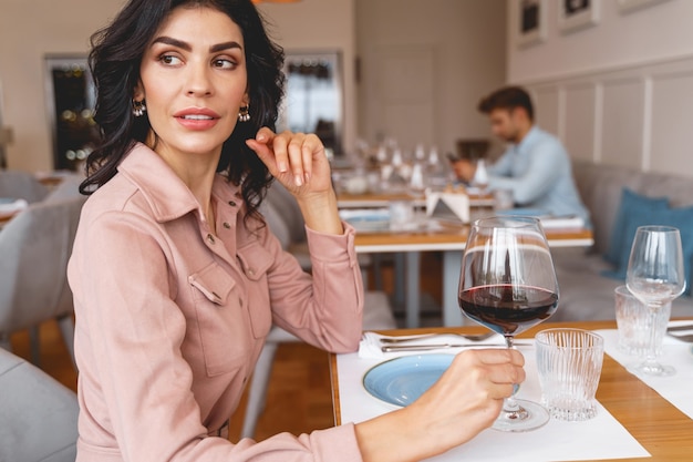 Foto bella giovane donna che tiene in mano un bicchiere di vino e distoglie lo sguardo mentre trascorre del tempo al ristorante