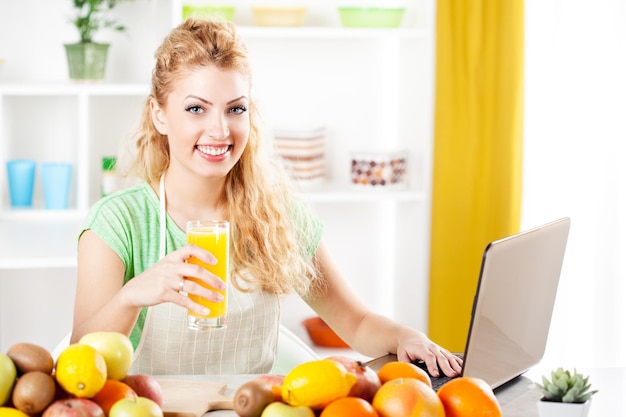 Bella giovane donna che tiene il succo e legge la ricetta con il computer portatile in una cucina. guardando la fotocamera.