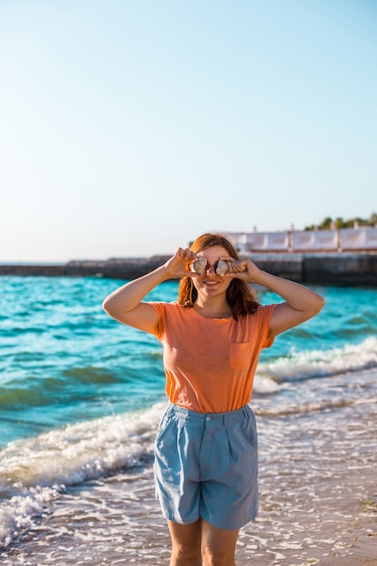 Bella giovane donna che tiene due conchiglie sul viso con gli occhi sulla spiaggia