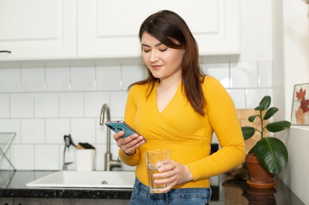 Beautiful young woman at home in the kitchen with a glass of water and a mobile phone