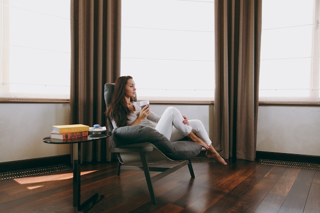 The beautiful young woman at home sitting on modern chair in front of window, relaxing in her living room and drinking coffee or tea