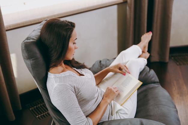 The beautiful young woman at home sitting on modern chair in front of window, relaxing in her living room and reading book