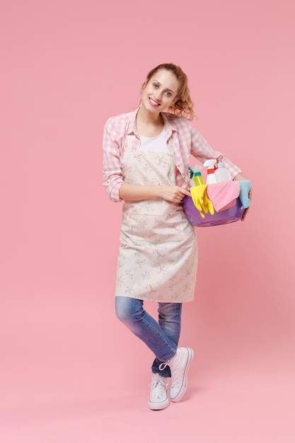 Beautiful young woman housewife in apron hold basin with detergent bottles washing cleansers doing housework isolated on pastel pink background studio portrait. Housekeeping concept. Looking camera.