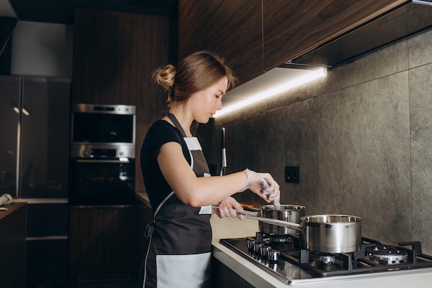 Красивая молодая женщина-домохозяйка готовит ужин, держит в руках большую стальную кастрюлю, стоящую на газовой плите
