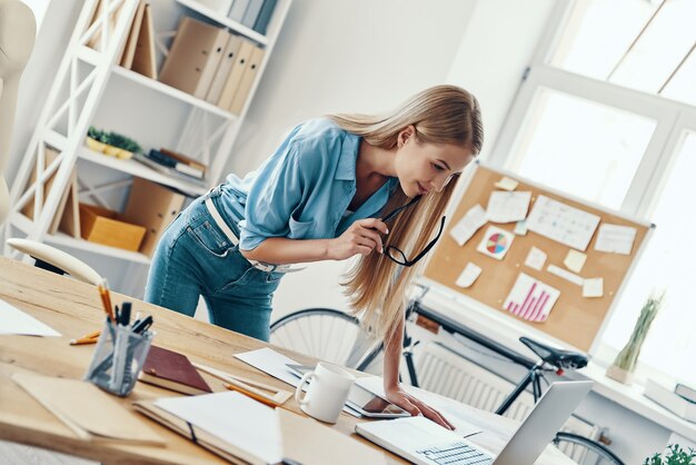 Фото Красивая молодая женщина в элегантной повседневной одежде улыбается и использует ноутбук во время работы в офисе