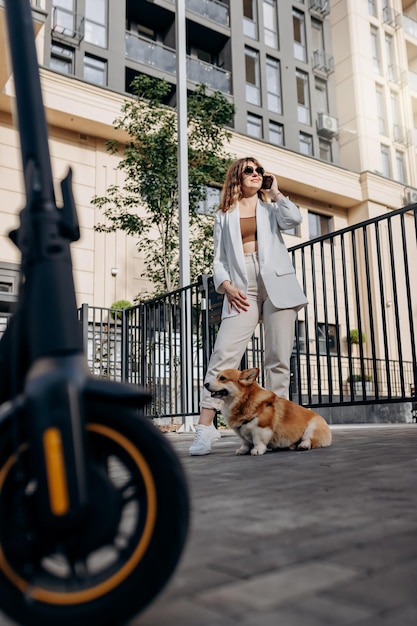 Фото Красивая молодая женщина в солнечных очках и белом костюме стоит со своим электрическим скутером и собакой корги возле современного здания в городе и разговаривает по телефону