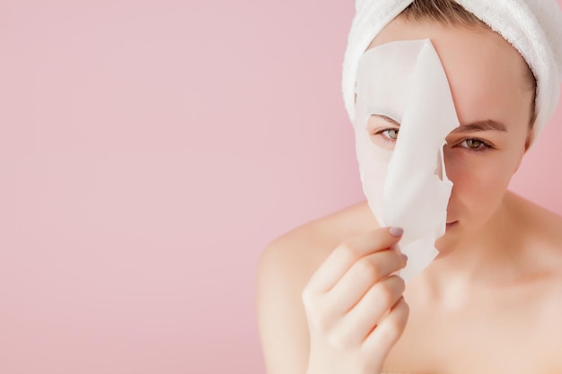 Bella giovane donna sta applicando una maschera cosmetica in tessuto su un viso su sfondo rosa trattamento sanitario e di bellezza e concetto tecnologico