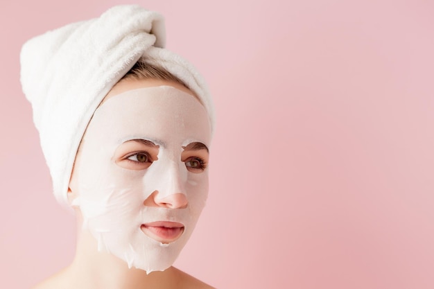 Beautiful young woman is applying a cosmetic tissue mask on a face on a pink background