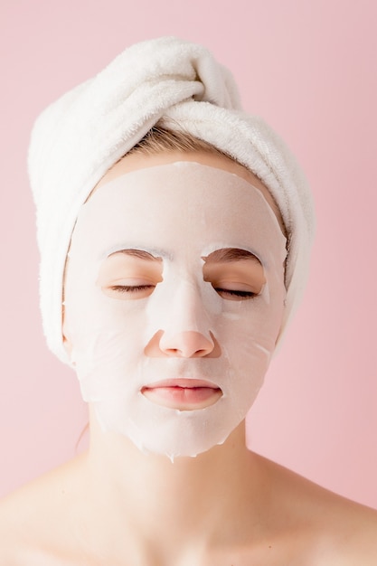 Beautiful young woman is applying a cosmetic tissue mask on a face on a pink.