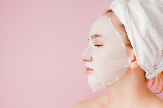 Beautiful young woman is applying a cosmetic tissue mask on a face on a pink