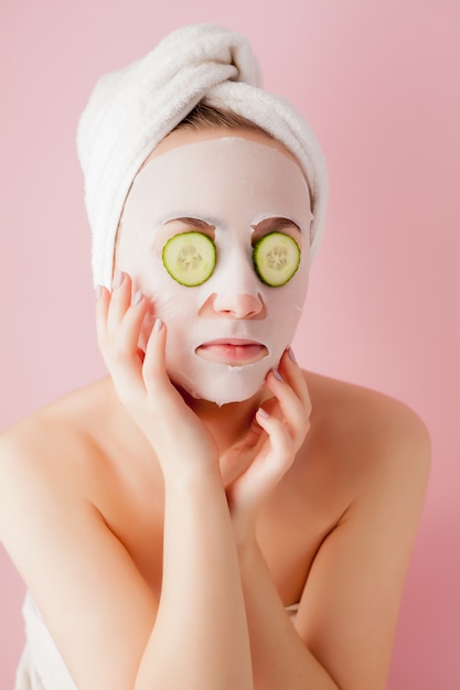 Beautiful young woman is applying a cosmetic tissue mask on a face with cucumber on a pink