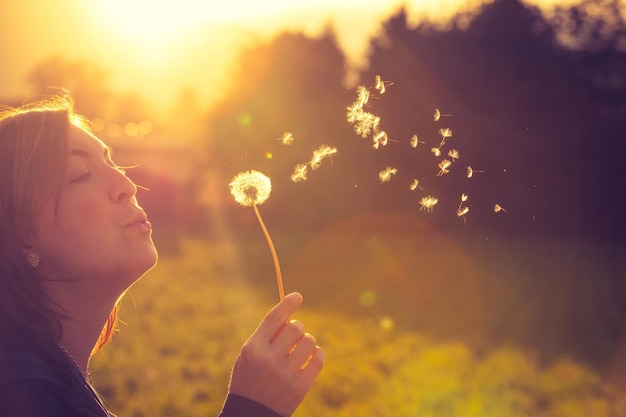 Beautiful young woman is blowing dandelion evening sun