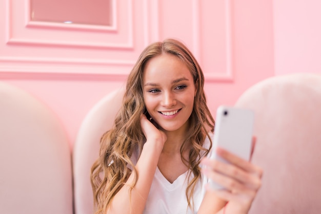 La bella giovane donna sta facendo selfie utilizzando uno smart phone, mostrando il segno giusto e sorridendo mentre riposa nella caffetteria