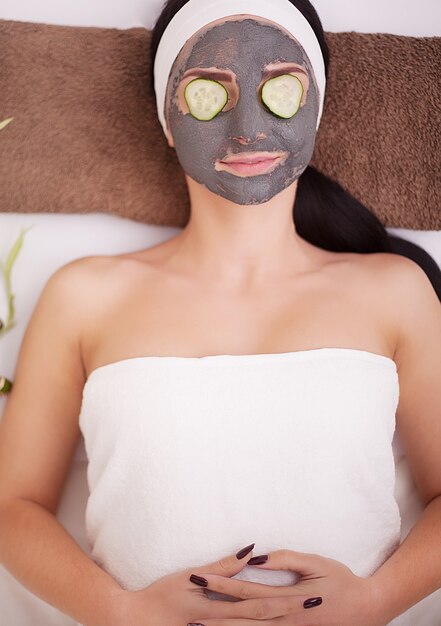 Beautiful young woman is getting facial clay mask at spa, lying with cucumbers on eyes