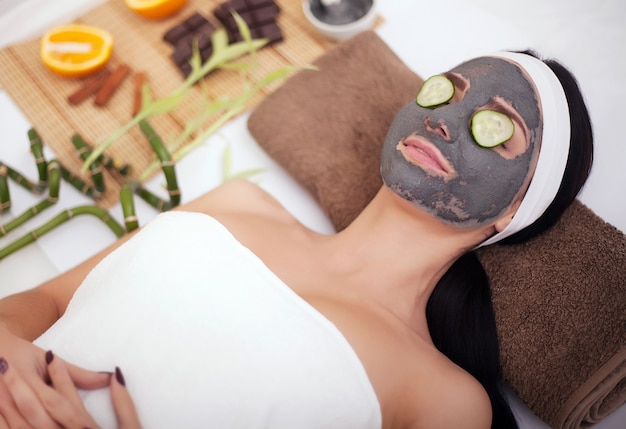 Beautiful young woman is getting facial clay mask at spa, lying with cucumbers on eyes