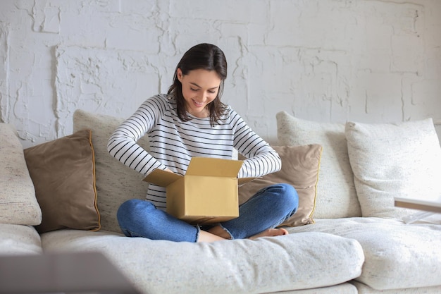 Красивая молодая женщина держит картонную коробку и распаковывает ее, сидя дома на диване