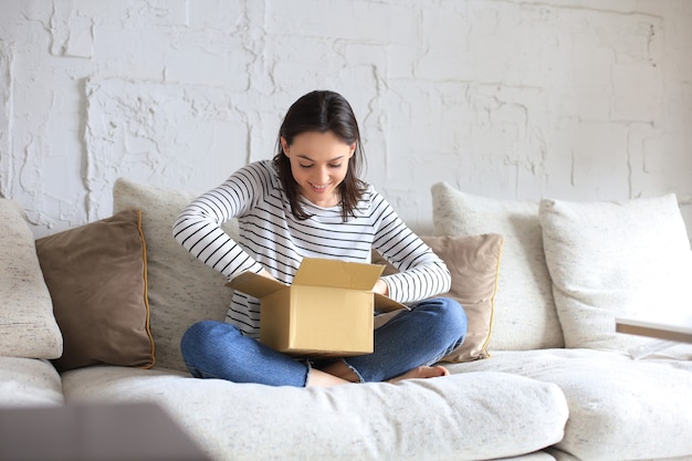 Красивая молодая женщина держит картонную коробку и распаковывает ее, сидя на диване у себя дома.
