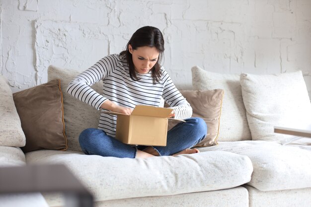 Beautiful young woman is holding cardboard box and unpacking it sitting on sofa at home.