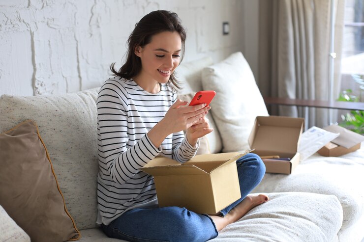 Photo beautiful young woman is holding cardboard box and unpacking smartphone sitting on sofa at home.