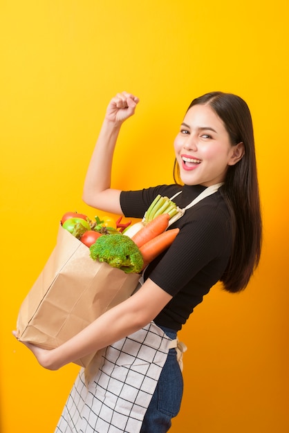 食料品の袋の黄色の壁に美しい若い女性が野菜を持っています。