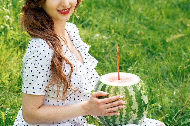 Beautiful young woman is holding a whole watermelon with cocktail straw sitting on grass, outdoors.