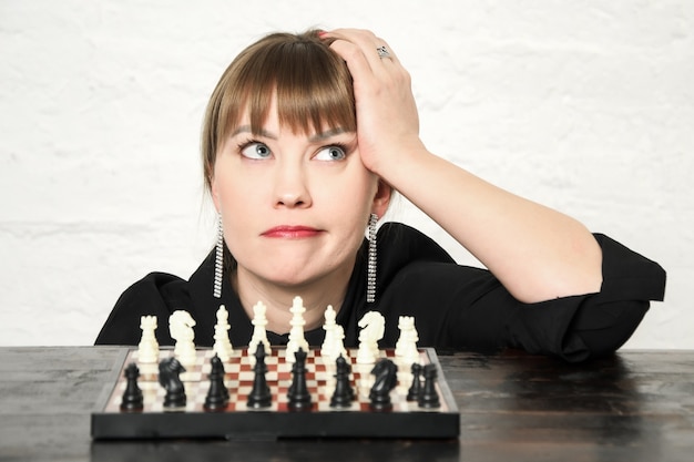 Фото Красивая молодая женщина задумчиво смотрит вверх, сидя перед шахматной доской с разнесенными фигурами