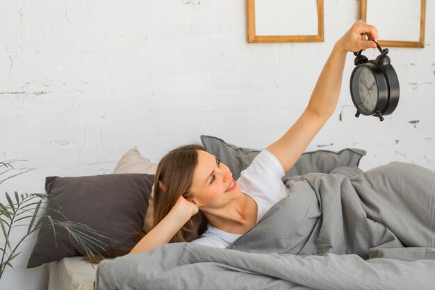 beautiful young woman is lying in bed under a blanket with an alarm clock in her hands