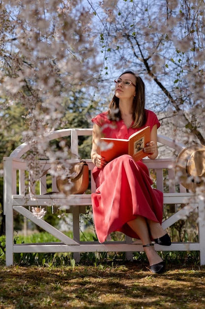 A beautiful young woman is sitting on an elegant bench in a spring garden under a cherry blossom and reading a book