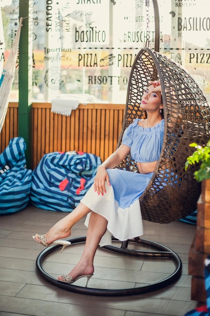 A beautiful young woman is sitting in a wicker hanging chair on the summer terrace of a city cafe looking at the camera and smiling