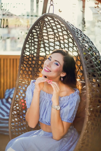 A beautiful young woman is sitting in a wicker hanging chair on the summer terrace of a city cafe she raised her hands to chin looking at the camera and smiling