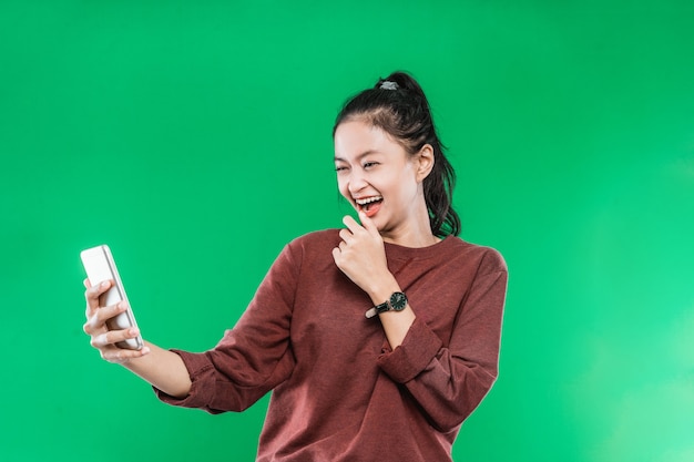 A beautiful young woman is on a video call with someone holding her chin face and laughing with a happy expression on a green background