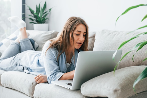 Beautiful young woman in jeans and blue shirt lying down at sofa with laptop