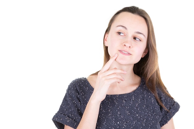 Beautiful young woman keeps finger on face looks aside with bewilderment poses against white background