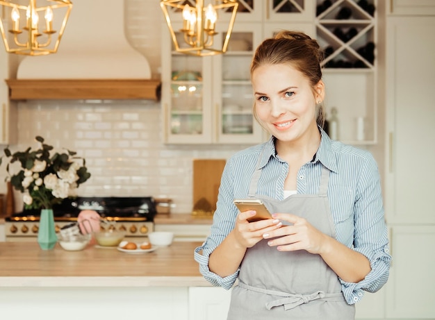 A beautiful young woman in the kitchen holds a smartphone and types a message for clients