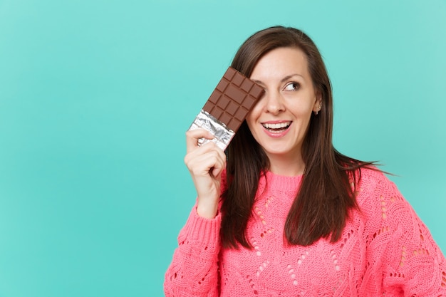 Beautiful young woman in knitted pink sweater looking aside hold in hand covering eye with chocolate bar isolated on blue wall background studio portrait. People lifestyle concept. Mock up copy space.