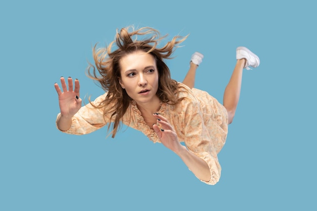 Photo beautiful young woman levitating in midair falling down and her hair messed up soaring from wind model flying hovering with dreamy peaceful expression indoor shot isolated on blue background