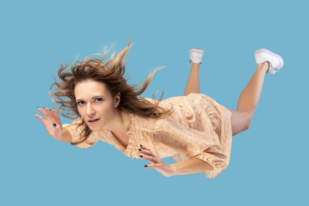 Beautiful young woman levitating in midair falling down and her hair messed up soaring from wind model flying hovering with dreamy peaceful expression indoor shot isolated on blue background