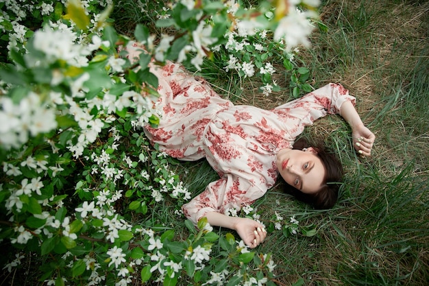 Beautiful young woman lies in grass under blooming apple tree. Romantic girl is resting in nature. Spring portrait of woman