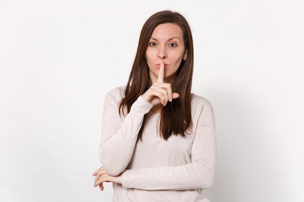 Beautiful young woman in light clothes saying hush be quiet with finger on lips shhh gesture isolated on white wall background in studio. People sincere emotions lifestyle concept. Mock up copy space.