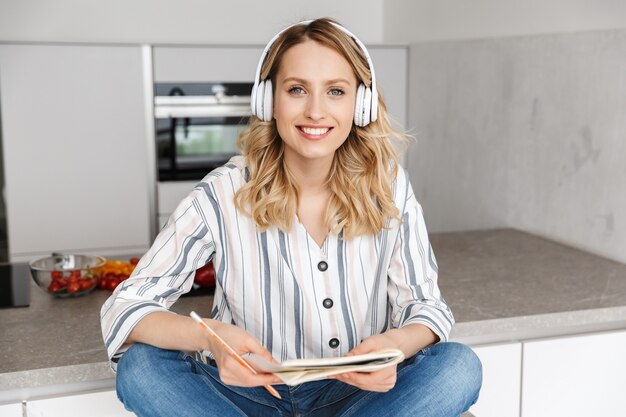 Красивая молодая женщина слушает музыку в наушниках, сидя на кухне, делая заметки