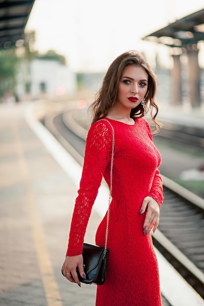 Beautiful young woman in a long red dress stands on the platform of the station near the railway
