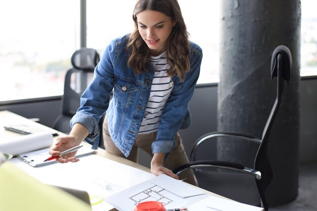 Beautiful young woman looking at blueprint and smiling while working in the creative office.