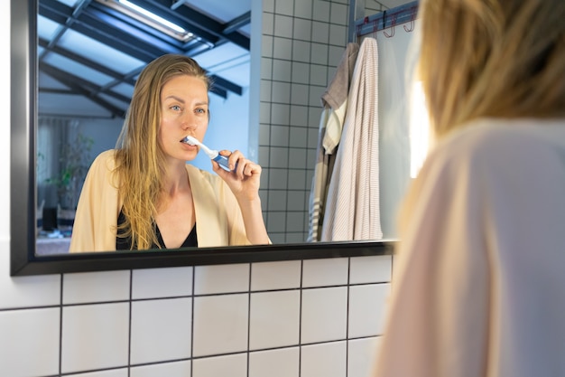 Bella giovane donna che esamina lo specchio del bagno lavarsi i denti con lo spazzolino da denti