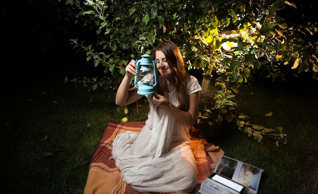 Beautiful young woman looking at old lantern while sitting at night garden