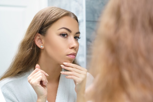 Beautiful young woman looks at her face in the mirror