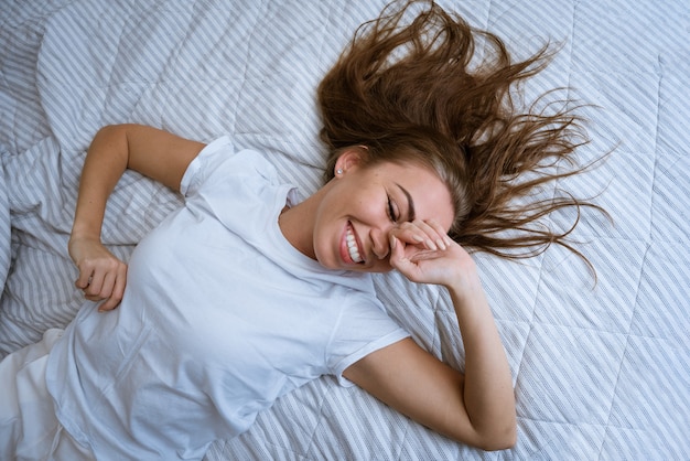 Beautiful young woman lying in bed smiling a girl with open eyes covers her face with her hands and ...